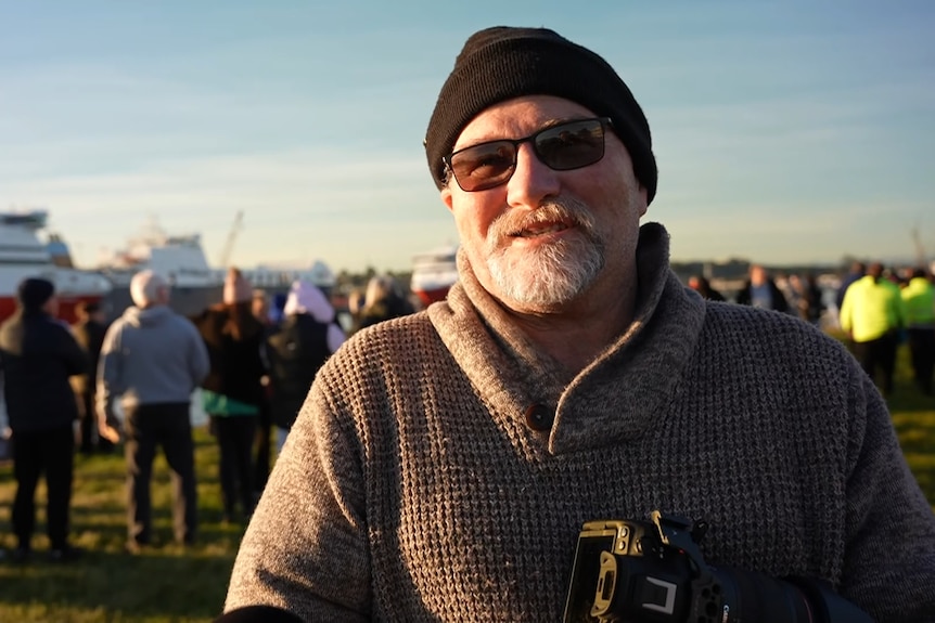 Rodney Winwood wears a woollen jumper and a beanie and holds a camera.