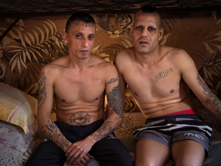 Two tattooed men sit shirtless on a bed under a bunk, looking directly at the camera in a dimly lit room.
