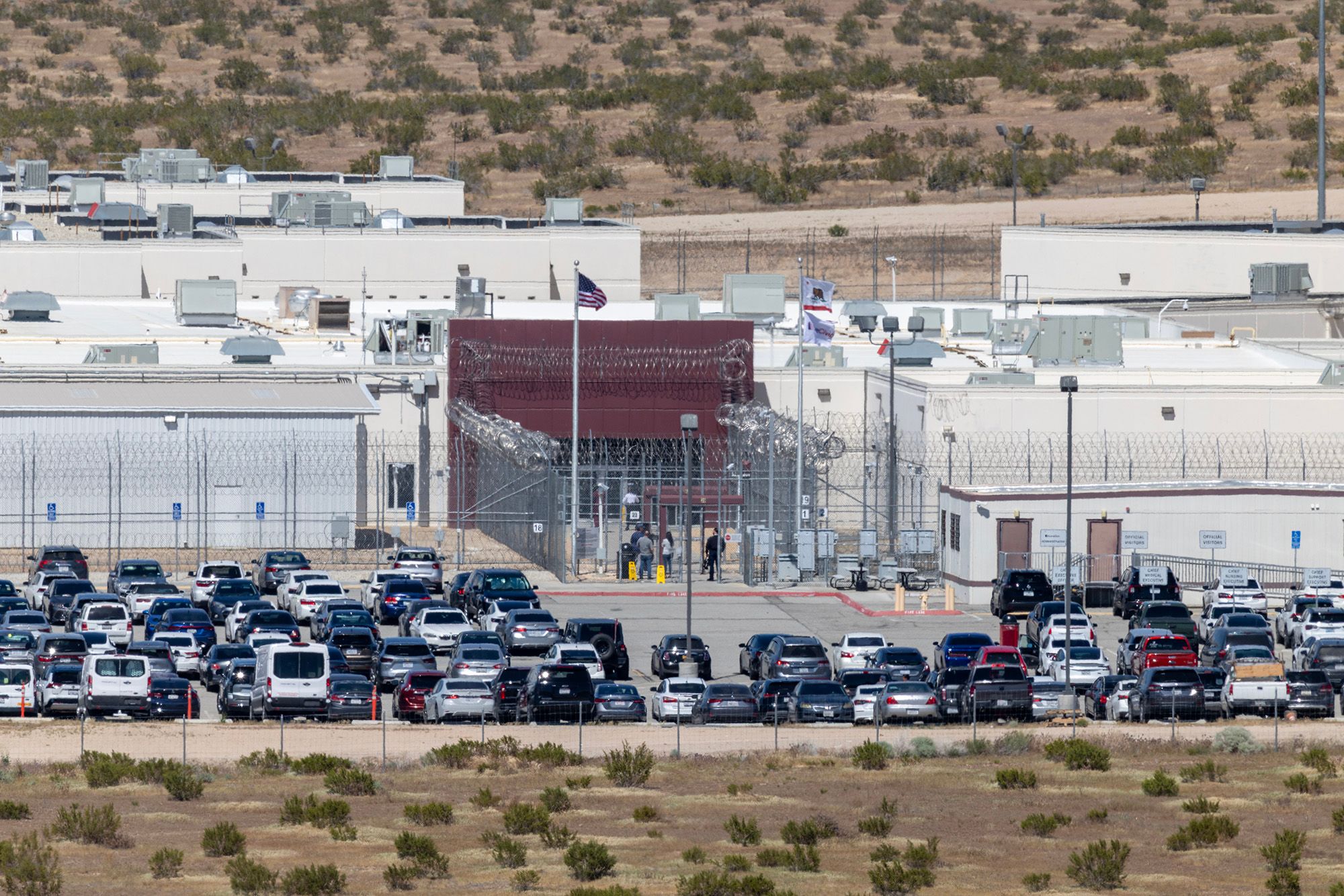 A photo shows the entrance of a detention facility surrounded by barbed wire fencing. Cars are parked in front of the building. 