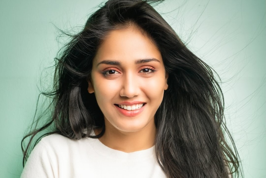 Twisha smiles, has loose dark hair flowing in front, wears white top.
