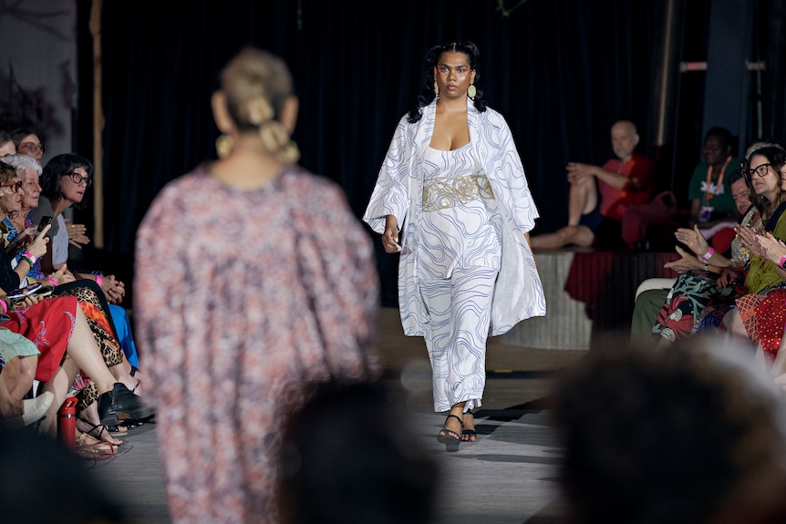 A model in a white outfit walking down a runway, at a night-time fashion show.