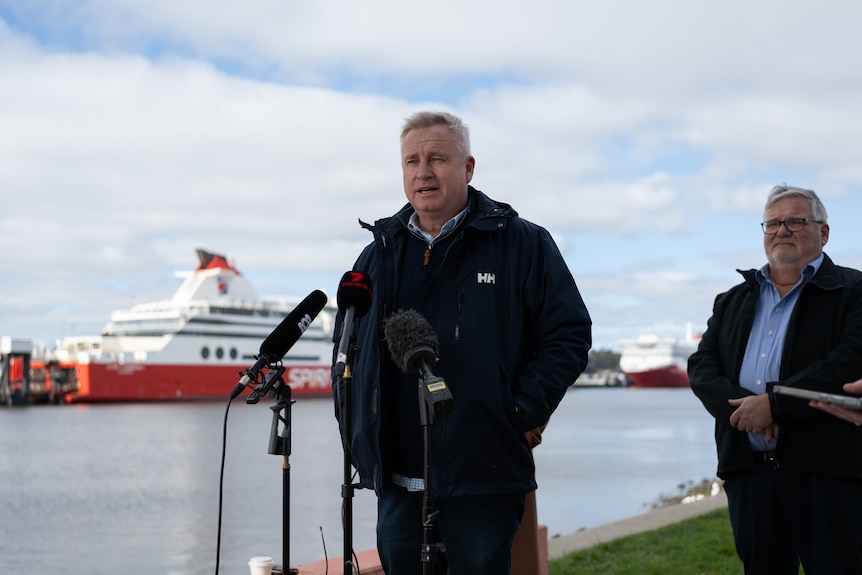 Jeremy Rockliff stands in front of microphones during press conference, with Mersey River and Spirit of Tasmania Ferries in t