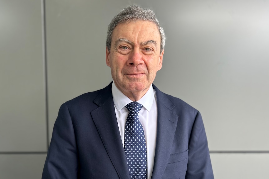 A grey-haired man in a blue suit and tie looks at the camera