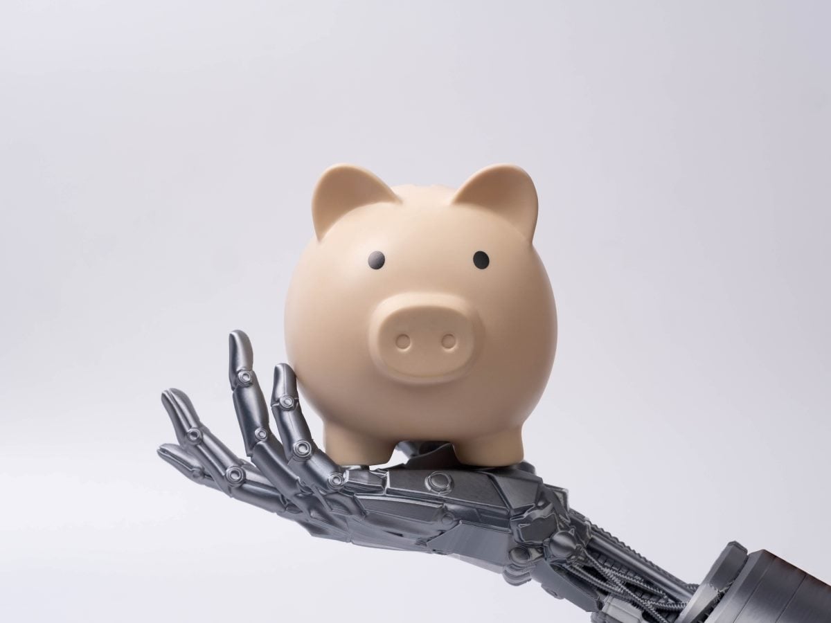 A beige piggy bank rests on the open palm of a metallic robotic hand, symbolizing the intersection of artificial intelligence, automation and financial management. The minimalist composition is set against a clean, light gray background with ample copy space.