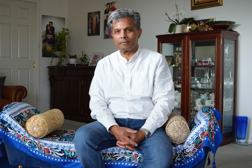 A man sits on a blue sofa in a living room in a crisp white shirt and looks at the camera.