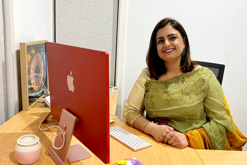 Reena, a smiling Indian woman sits at desk in front of Apple monitor, wears traditional clothes, green scarf across bust.
