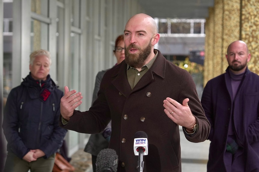 A man with a beard speaks animatedly in front of a microphone.