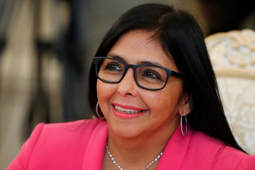 Delcy Rodriguez smiling, wearing a bright pink jacket, silver necklace and dark-rimmed glasses.
