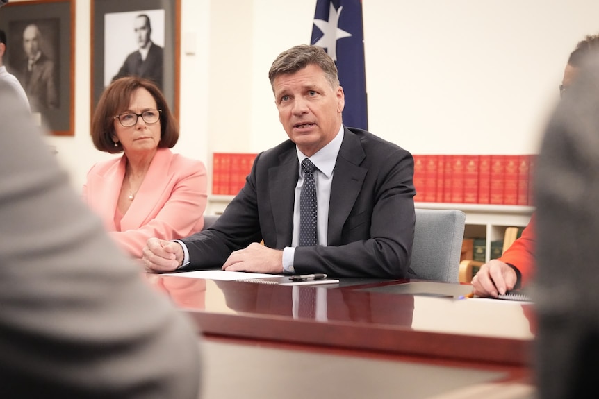 Man in white shirt and dark suit and tie sitting at long table and speaking with Australian flag in background.