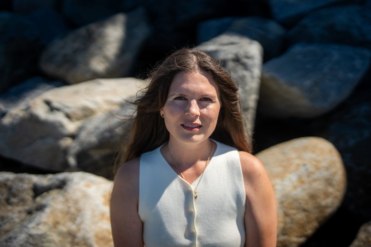 A woman with long brown hair sits on a rock looking directly into the camera.
