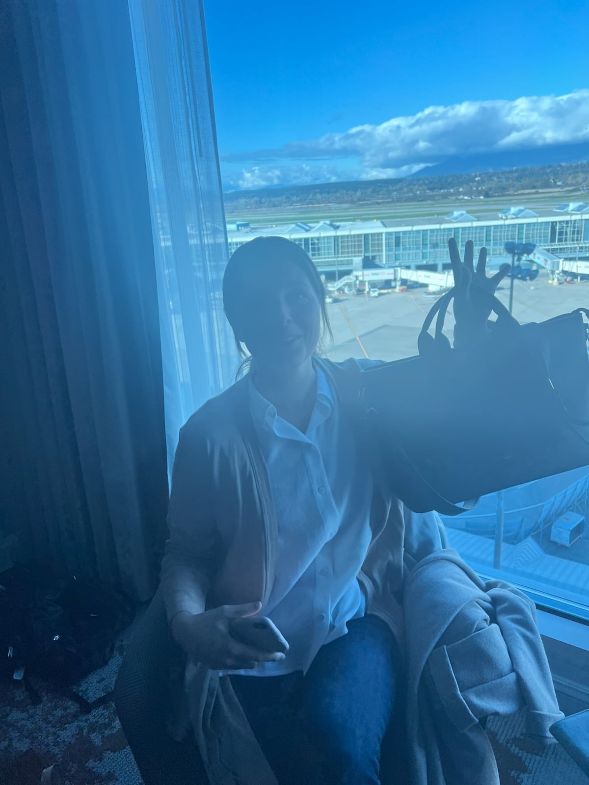 A brunette woman with a white blouse and a light brown sweater sits in front of a window holding up a large purse with an airport visible through the window.