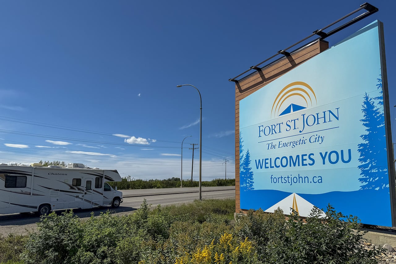 A welcome to Fort St. John sign can be seen along the Alaska Highway. A white RV is driving from right to left along the highway toward the city.