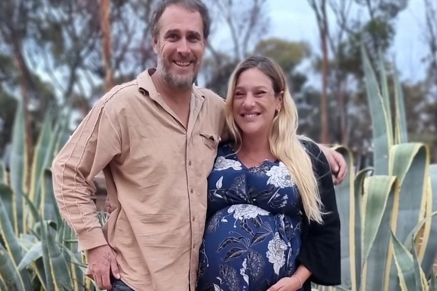 Man in a brown shirt stands next a blonde pregnant woman in a blue dress. In front of them is a young blonde boy