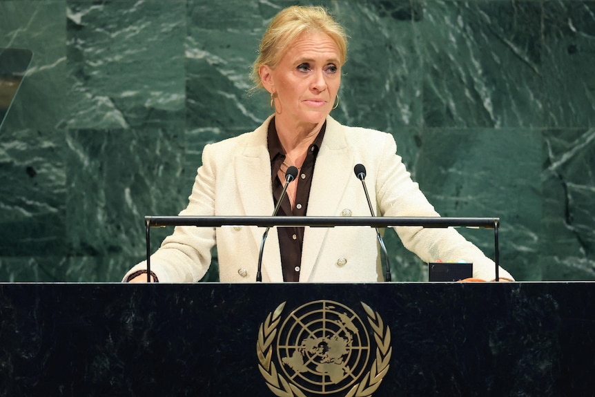 Þorgerður Katrín Gunnarsdóttir in a white blazer and brown top speaking at a black UN lectern in front of a green wall.