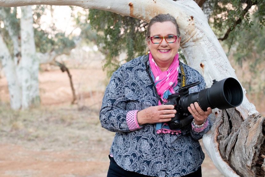 Woman holding camera, wearing a black and white shirt and pink scarf, stands smiling in front of trees in the dry outback