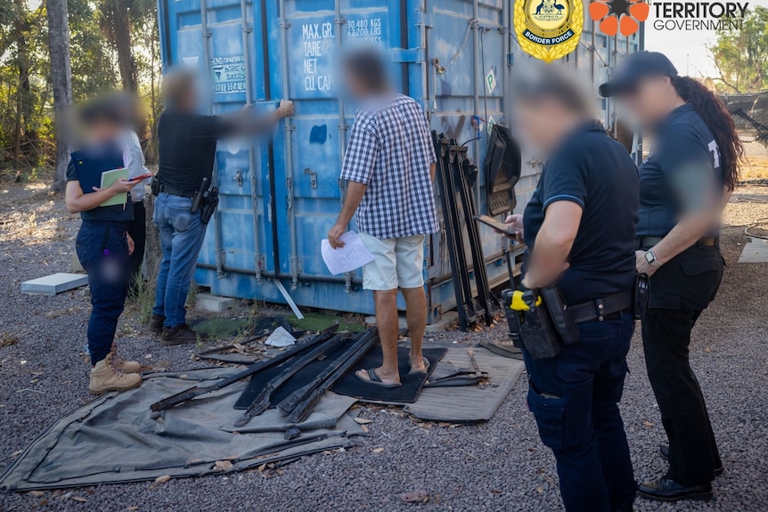 Multiple law enforcement officers prepare to open a blue shipping container while a man in civilian clothing looks on.